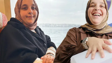 Görselde başörtülü iki kadın yan yana oturuyor. Soldaki kadın kameraya bakarken, sağdaki kadın gülümseyerek elini önündeki beyaz kutunun üzerine koyuyor. Arka planda deniz manzarası görülüyor.