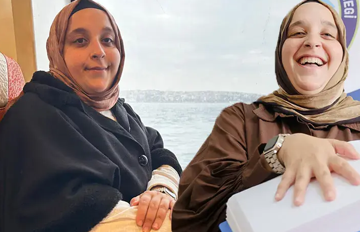 Görselde başörtülü iki kadın yan yana oturuyor. Soldaki kadın kameraya bakarken, sağdaki kadın gülümseyerek elini önündeki beyaz kutunun üzerine koyuyor. Arka planda deniz manzarası görülüyor.