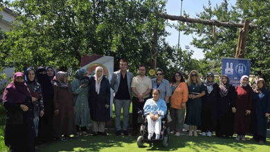 Fotoğrafta, açık havada düzenlenen etkinlikte engelli anneleri ve katılımcılar toplu halde objektife poz veriyor. Ön sırada tekerlekli sandalye kullanan bir kişi yer alırken, arka planda ağaçlar, yeşil alan ve etkinliğe ait afişler görülüyor.