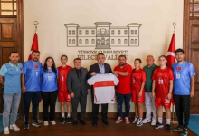 Fotoğrafta, İşitme Engelliler Genç Kız Futsal Milli Takımı sporcuları, antrenörleri ve yetkililer Bilecik Valiliği binasında toplu halde poz veriyor. Ortada yer alan yetkililer milli takım formasını birlikte tutarken, sporcular kırmızı ve mavi milli takım formalarıyla iki tarafta sıralanıyor. Arka planda Bilecik Valiliği yazısı ve iki Türk bayrağı yer alıyor. Haber, İşitme Engelliler Genç Kız Futsal Milli Takımı'nın uluslararası organizasyon hazırlıkları kapsamında Bilecik'te kampa girmesini konu alıyor.
