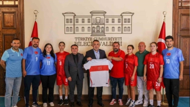 Fotoğrafta, İşitme Engelliler Genç Kız Futsal Milli Takımı sporcuları, antrenörleri ve yetkililer Bilecik Valiliği binasında toplu halde poz veriyor. Ortada yer alan yetkililer milli takım formasını birlikte tutarken, sporcular kırmızı ve mavi milli takım formalarıyla iki tarafta sıralanıyor. Arka planda Bilecik Valiliği yazısı ve iki Türk bayrağı yer alıyor. Haber, İşitme Engelliler Genç Kız Futsal Milli Takımı'nın uluslararası organizasyon hazırlıkları kapsamında Bilecik'te kampa girmesini konu alıyor.