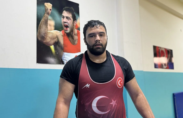 Fotoğrafta, işitme engelli milli güreşçi Berke Nergiz antrenman salonunda görülüyor. Nergiz, üzerinde Türk bayrağı ve ay-yıldız bulunan kırmızı güreş mayosuyla kameraya poz veriyor. Arka plandaki duvarda bir güreşçinin müsabaka sırasında çekilmiş büyük bir fotoğrafı yer alıyor. Haber, Berke Nergiz'in Avrupa'da başarı elde etme hedefini konu alıyor.
