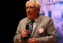Fotoğrafta KASDER'in Kurucusu ve Onursal Başkanı Prof. Dr. Coşkun Özdemir, takım elbisesiyle sahnede mikrofonla konuşurken görülüyor.