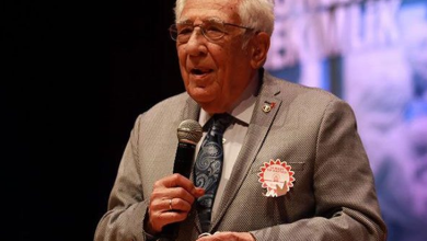 Fotoğrafta KASDER'in Kurucusu ve Onursal Başkanı Prof. Dr. Coşkun Özdemir, takım elbisesiyle sahnede mikrofonla konuşurken görülüyor.