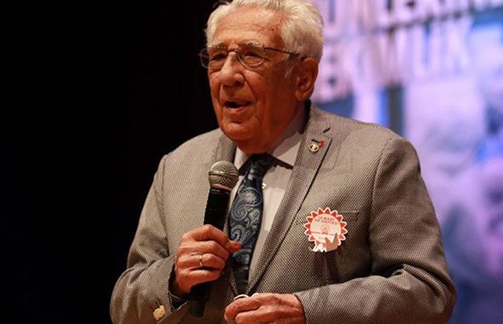 Fotoğrafta KASDER'in Kurucusu ve Onursal Başkanı Prof. Dr. Coşkun Özdemir, takım elbisesiyle sahnede mikrofonla konuşurken görülüyor.