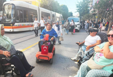 Fotoğrafta akülü ve manuel tekerlekli sandalye kullanan bireyler, tramvay hattının bulunduğu kalabalık bir caddede bir araya geliyor. Arka planda tramvay, yayalar ve şehir trafiği görülürken, katılımcılar sohbet ediyor.
