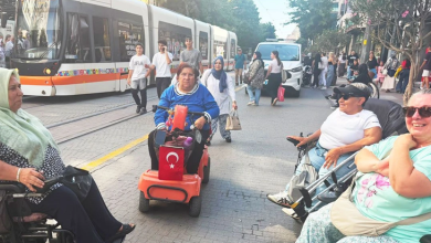Fotoğrafta akülü ve manuel tekerlekli sandalye kullanan bireyler, tramvay hattının bulunduğu kalabalık bir caddede bir araya geliyor. Arka planda tramvay, yayalar ve şehir trafiği görülürken, katılımcılar sohbet ediyor.