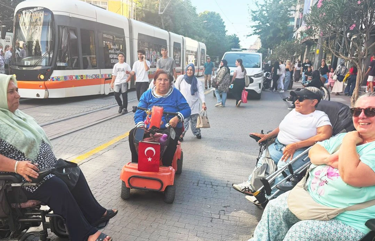Fotoğrafta akülü ve manuel tekerlekli sandalye kullanan bireyler, tramvay hattının bulunduğu kalabalık bir caddede bir araya geliyor. Arka planda tramvay, yayalar ve şehir trafiği görülürken, katılımcılar sohbet ediyor.