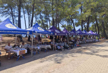 Fotoğrafta, Muğla Büyükşehir Belediyesine ait çadırların kurulduğu açık alanda engelli bireyler ve ailelerinin el emeği ürünlerini sergilediği stantlar görülüyor. Ağaçlık alanda kurulan stantlarda ziyaretçiler alışveriş yapıyor ve ürünleri inceliyor.