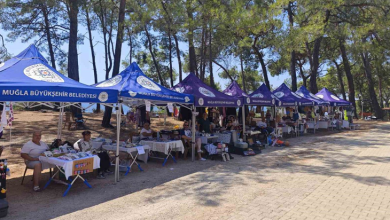 Fotoğrafta, Muğla Büyükşehir Belediyesine ait çadırların kurulduğu açık alanda engelli bireyler ve ailelerinin el emeği ürünlerini sergilediği stantlar görülüyor. Ağaçlık alanda kurulan stantlarda ziyaretçiler alışveriş yapıyor ve ürünleri inceliyor.