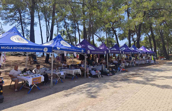 Fotoğrafta, Muğla Büyükşehir Belediyesine ait çadırların kurulduğu açık alanda engelli bireyler ve ailelerinin el emeği ürünlerini sergilediği stantlar görülüyor. Ağaçlık alanda kurulan stantlarda ziyaretçiler alışveriş yapıyor ve ürünleri inceliyor.