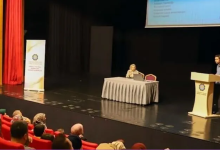 Konferans salonunda düzenlenen bir toplantıda katılımcılar kırmızı koltuklarda otururken, sahnede kürsüden sunum yapan bir konuşmacı ve masada oturan bir moderatör görülüyor. Arka planda projeksiyon perdesi ve etkinlik afişi yer alırken, izleyiciler sunumu dikkatle takip ediyor.