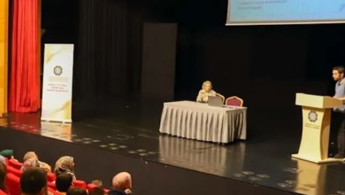 Konferans salonunda düzenlenen bir toplantıda katılımcılar kırmızı koltuklarda otururken, sahnede kürsüden sunum yapan bir konuşmacı ve masada oturan bir moderatör görülüyor. Arka planda projeksiyon perdesi ve etkinlik afişi yer alırken, izleyiciler sunumu dikkatle takip ediyor.