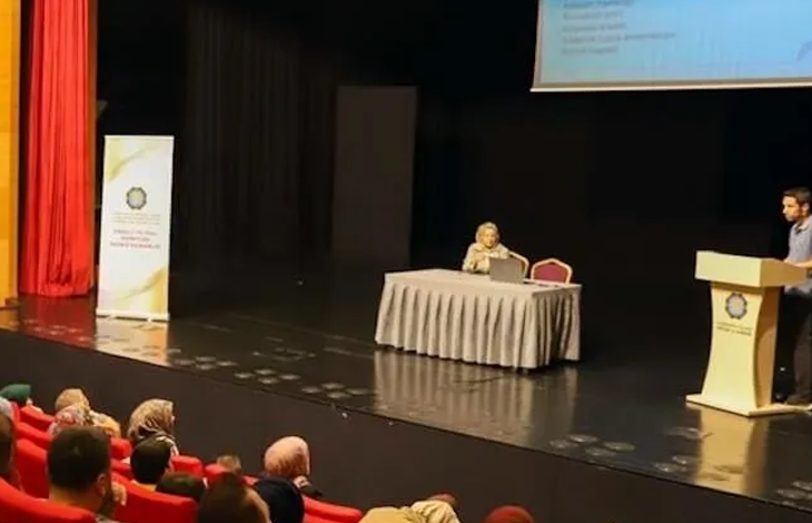 Konferans salonunda düzenlenen bir toplantıda katılımcılar kırmızı koltuklarda otururken, sahnede kürsüden sunum yapan bir konuşmacı ve masada oturan bir moderatör görülüyor. Arka planda projeksiyon perdesi ve etkinlik afişi yer alırken, izleyiciler sunumu dikkatle takip ediyor.