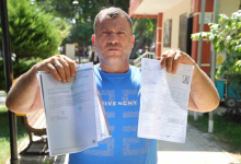 Fotoğrafta, açık mavi tişört giyen bir kişi elinde resmi belgeleri tutarak kameraya gösteriyor. Açık alanda çekilen fotoğrafın arka planında ağaçlar ve bir kamu binası görülüyor.