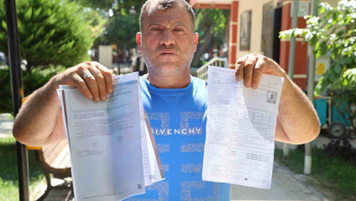Fotoğrafta, açık mavi tişört giyen bir kişi elinde resmi belgeleri tutarak kameraya gösteriyor. Açık alanda çekilen fotoğrafın arka planında ağaçlar ve bir kamu binası görülüyor.
