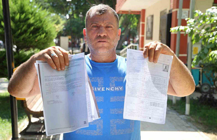 Fotoğrafta, açık mavi tişört giyen bir kişi elinde resmi belgeleri tutarak kameraya gösteriyor. Açık alanda çekilen fotoğrafın arka planında ağaçlar ve bir kamu binası görülüyor.