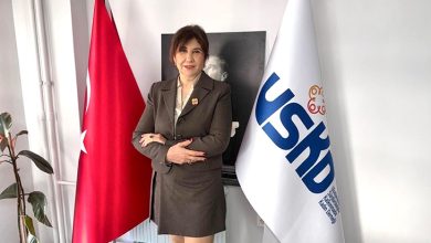 Resimde, koyu renk bir blazer ve etek takımı giyen Ayla Alkan, kolları bağlı şekilde kendinden emin bir duruşla ayakta görülüyor. Arka planda Mustafa Kemal Atatürk’ün siyah-beyaz portresi yer alıyor. Sağında USKD bayrağı, solunda ise Türk bayrağı bulunuyor. Ortam, kurumsal bir ofis veya dernek çalışma alanı izlenimi veriyor.