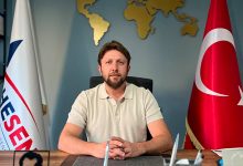 Fotoğrafta Dr. Ahmet Kandemir, çalışma masasında otururken görülüyor. Arkasında Türkiye bayrağı ve AHESEN bayrağı bulunuyor. Duvarında altın tonlarında dekoratif bir dünya haritası yer alıyor. Masada evraklar, kalemlik ve Atatürk silueti bulunan bir obje dikkat çekiyor. Ortam resmi ve kurumsal bir ofis atmosferi yansıtıyor.