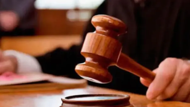 Görselde, bir kişinin elinde tuttuğu ahşap hâkim tokmağı ve masadaki tokmak altlığı yakın planda görülüyor. Arka planda duruşma ortamını çağrıştıran bulanık kişiler yer alıyor.