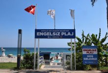 Görselde, deniz kenarında Antalya Büyükşehir Belediyesi’ne ait “Engelsiz Plaj” girişinin, Türk bayrağı ve çeşitli bayraklarla süslenmiş şekilde, erişilebilir rampalar ve yürüyüş yollarıyla birlikte gösterildiği görülüyor.