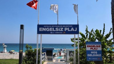 Görselde, deniz kenarında Antalya Büyükşehir Belediyesi’ne ait “Engelsiz Plaj” girişinin, Türk bayrağı ve çeşitli bayraklarla süslenmiş şekilde, erişilebilir rampalar ve yürüyüş yollarıyla birlikte gösterildiği görülüyor.