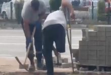 Engelli personel değneğini tutarak, kaldırım döşemek için kazma kullanan personele yardım ediyor.