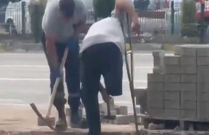 Engelli personel değneğini tutarak, kaldırım döşemek için kazma kullanan personele yardım ediyor.
