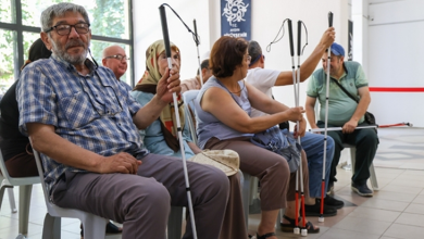 Bir salonda yan yana oturan görme engelli bireyler, ellerinde farklı türlerde beyaz bastonlar tutarken görülüyor.