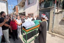 Görselde, kalabalık bir grubun cenaze töreni sırasında tabutun başında toplandığı ve önde bir din görevlisinin dua ettiği görülüyor.