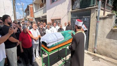 Görselde, kalabalık bir grubun cenaze töreni sırasında tabutun başında toplandığı ve önde bir din görevlisinin dua ettiği görülüyor.