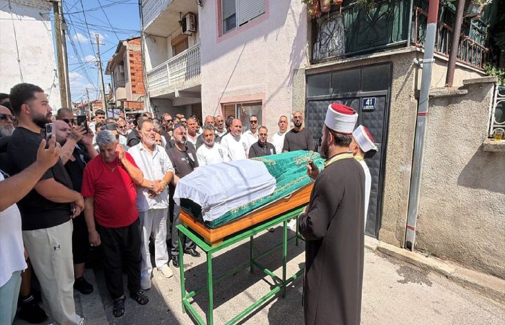 Görselde, kalabalık bir grubun cenaze töreni sırasında tabutun başında toplandığı ve önde bir din görevlisinin dua ettiği görülüyor.