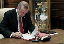 Fotoğrafta Cumhurbaşkanı Recep Tayyip Erdoğan, çalışma masasında resmi evrakları inceleyip imzalarken görülüyor. Üzerinde koyu renk takım elbise, beyaz gömlek ve kırmızı kravat bulunuyor. Masada kırmızı bir dosya, kalem, mühür ve çeşitli büro malzemeleri yer alıyor. Arka planda camlı vitrinde altın renkli ödül veya hatıra objeleri sergileniyor. Ortam, Cumhurbaşkanlığı makam odası niteliğinde resmi bir çalışma atmosferi taşıyor.