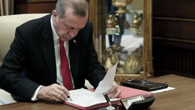 Fotoğrafta Cumhurbaşkanı Recep Tayyip Erdoğan, çalışma masasında resmi evrakları inceleyip imzalarken görülüyor. Üzerinde koyu renk takım elbise, beyaz gömlek ve kırmızı kravat bulunuyor. Masada kırmızı bir dosya, kalem, mühür ve çeşitli büro malzemeleri yer alıyor. Arka planda camlı vitrinde altın renkli ödül veya hatıra objeleri sergileniyor. Ortam, Cumhurbaşkanlığı makam odası niteliğinde resmi bir çalışma atmosferi taşıyor.