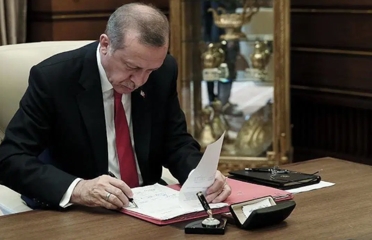 Fotoğrafta Cumhurbaşkanı Recep Tayyip Erdoğan, çalışma masasında resmi evrakları inceleyip imzalarken görülüyor. Üzerinde koyu renk takım elbise, beyaz gömlek ve kırmızı kravat bulunuyor. Masada kırmızı bir dosya, kalem, mühür ve çeşitli büro malzemeleri yer alıyor. Arka planda camlı vitrinde altın renkli ödül veya hatıra objeleri sergileniyor. Ortam, Cumhurbaşkanlığı makam odası niteliğinde resmi bir çalışma atmosferi taşıyor.