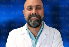 Fotoğrafta Doç. Dr. Ümit Alakuş, beyaz hekim önlüğü giymiş, sakallı ve gözlüklü bir erkek. Kollarını göğsünde birleştirmiş şekilde duruyor. Üzerindeki önlükte “Egepol Hastaneleri” logosu bulunuyor. Arka planda aynı logonun tekrarlandığı mavi bir tanıtım panosu var. Doktor, profesyonel ve güven veren bir ifadeyle gülümsüyor.
