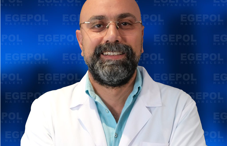Fotoğrafta Doç. Dr. Ümit Alakuş, beyaz hekim önlüğü giymiş, sakallı ve gözlüklü bir erkek. Kollarını göğsünde birleştirmiş şekilde duruyor. Üzerindeki önlükte “Egepol Hastaneleri” logosu bulunuyor. Arka planda aynı logonun tekrarlandığı mavi bir tanıtım panosu var. Doktor, profesyonel ve güven veren bir ifadeyle gülümsüyor.