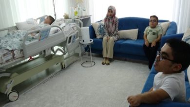Fotoğrafta hastane yatağında yatan bir engelli çocuk, yanında oturan iki çocuk ve karşıdaki koltukta oturan bir anne Sevim Akın görülüyor; odada tıbbi cihazlar ve yatak çevresinde çeşitli sağlık ekipmanları bulunuyor.