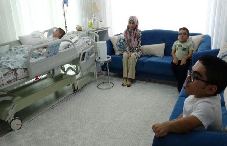 Fotoğrafta hastane yatağında yatan bir engelli çocuk, yanında oturan iki çocuk ve karşıdaki koltukta oturan bir anne Sevim Akın görülüyor; odada tıbbi cihazlar ve yatak çevresinde çeşitli sağlık ekipmanları bulunuyor.