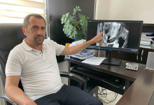 Bir ofis ortamında, masasında oturan Enver Gökçe görülüyor. Önünde bilgisayar klavyesi, hesap makinesi, evraklar ve çalışma malzemeleri bulunan düzenli bir masa var. Enver Gökçe sağ eliyle bilgisayar monitörünü işaret ediyor. Monitörde halk otobüsüne ait güvenlik kamerası görüntüleri açık; otobüs içini gösteren siyah‑beyaz kayıtlar ekranda belirgin şekilde görülüyor. Arka planda bir pencere ve yanında küçük bir bitki bulunuyor. Ortam genel olarak sakin, profesyonel ve denetim/izleme amaçlı bir çalışma alanı izlenimi veriyor.