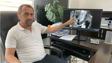 Bir ofis ortamında, masasında oturan Enver Gökçe görülüyor. Önünde bilgisayar klavyesi, hesap makinesi, evraklar ve çalışma malzemeleri bulunan düzenli bir masa var. Enver Gökçe sağ eliyle bilgisayar monitörünü işaret ediyor. Monitörde halk otobüsüne ait güvenlik kamerası görüntüleri açık; otobüs içini gösteren siyah‑beyaz kayıtlar ekranda belirgin şekilde görülüyor. Arka planda bir pencere ve yanında küçük bir bitki bulunuyor. Ortam genel olarak sakin, profesyonel ve denetim/izleme amaçlı bir çalışma alanı izlenimi veriyor.