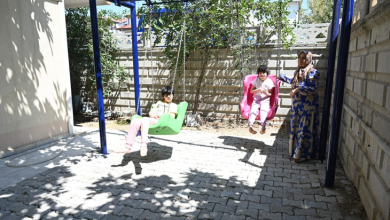 Bahçede kurulan salıncaklarda iki engelli kardeş otururken, yanlarında bir kadın ayakta duruyor.