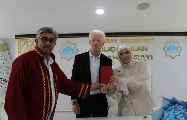 Görselde, nikâh töreninde görme engelli çift yan yana dururken nikâh görevlisiyle birlikte evlilik cüzdanını tutuyor. Arka planda Aksaray Belediyesi logosu ve nikâh salonuna ait yazılar görülüyor.