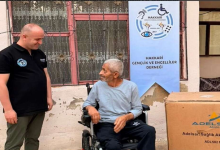 Görselde akülü tekerlekli sandalyede oturan bir kişi ile yanında duran bir görevli görülüyor. Arka planda Hakkari Gençlik ve Engelliler Derneği afişi, sağ tarafta ise tekerlekli sandalyeye ait büyük bir kutu yer alıyor.