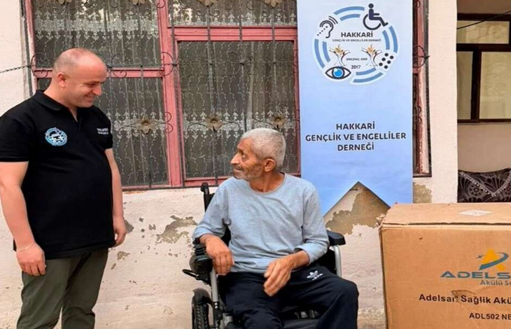 Görselde akülü tekerlekli sandalyede oturan bir kişi ile yanında duran bir görevli görülüyor. Arka planda Hakkari Gençlik ve Engelliler Derneği afişi, sağ tarafta ise tekerlekli sandalyeye ait büyük bir kutu yer alıyor.