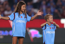 Görselde Trabzonspor forması giyen Isaac Kearney ile yanında yine Trabzonspor forması giyen bir kız çocuğu futbol sahasında görülüyor. Isaac Kearney elleriyle taraftarlara doğru işaret yaparken kız çocuğu kollarını iki yana açıyor. Arka planda tribünler yer alıyor.