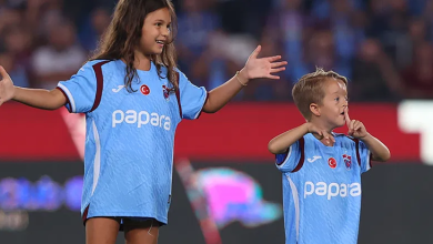 Görselde Trabzonspor forması giyen Isaac Kearney ile yanında yine Trabzonspor forması giyen bir kız çocuğu futbol sahasında görülüyor. Isaac Kearney elleriyle taraftarlara doğru işaret yaparken kız çocuğu kollarını iki yana açıyor. Arka planda tribünler yer alıyor.