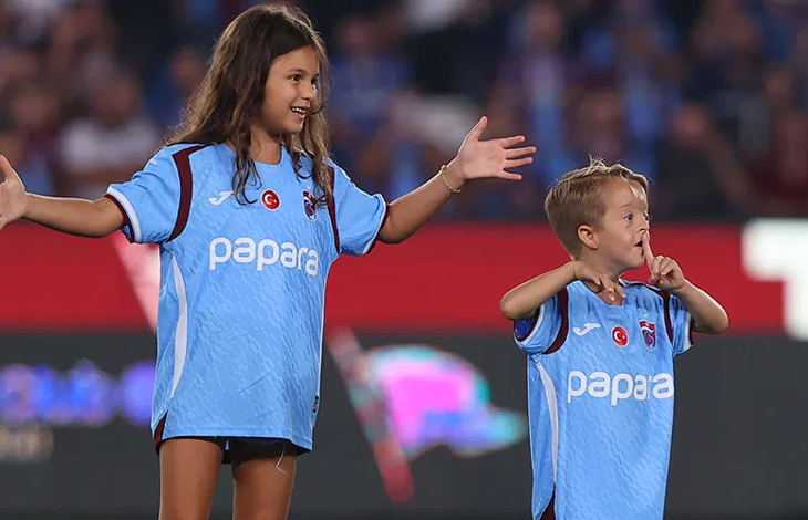 Görselde Trabzonspor forması giyen Isaac Kearney ile yanında yine Trabzonspor forması giyen bir kız çocuğu futbol sahasında görülüyor. Isaac Kearney elleriyle taraftarlara doğru işaret yaparken kız çocuğu kollarını iki yana açıyor. Arka planda tribünler yer alıyor.