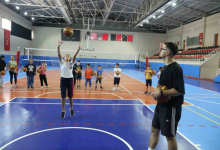 Kapalı spor salonunda çocuklar ellerinde basketbol toplarıyla sıraya girmiş durumda. Önde bir kadın potaya doğru top atarken, yanında elinde basketbol topu bulunan bir antrenör onu izliyor.