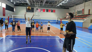 Kapalı spor salonunda çocuklar ellerinde basketbol toplarıyla sıraya girmiş durumda. Önde bir kadın potaya doğru top atarken, yanında elinde basketbol topu bulunan bir antrenör onu izliyor.